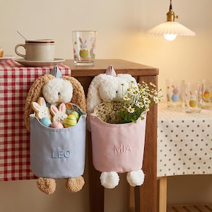 May include: Two plush bunny-shaped hanging baskets, one blue with "LEO" embroidered, the other pink with "MIA". The blue basket holds Easter cookies and eggs, the pink one, flowers. A glass with a bunny design is visible.