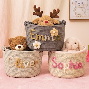 May include: Three round, woven storage baskets in gray, beige, and light beige, each with a child's name stitched on the side. The gray basket has the name "Emma" and a reindeer plush toy. The beige basket has the name "Oliver" and a teddy bear. The light beige basket has the name "Sophia" and a bunny.