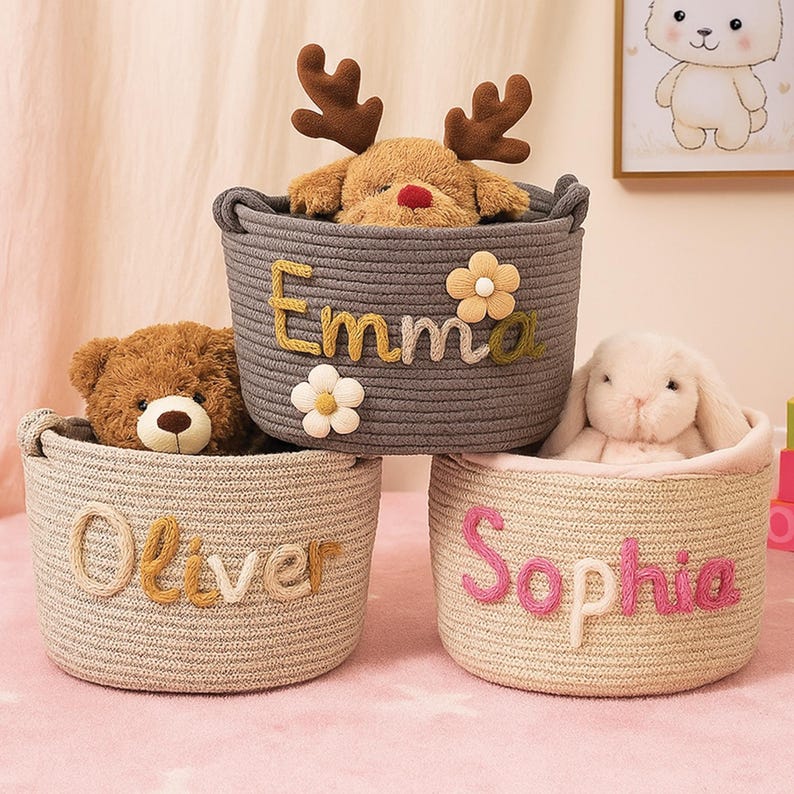 May include: Three round, woven storage baskets in gray, beige, and light beige, each with a child's name stitched on the side. The gray basket has the name "Emma" and a reindeer plush toy. The beige basket has the name "Oliver" and a teddy bear. The light beige basket has the name "Sophia" and a bunny.