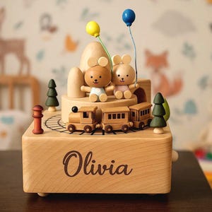 May include: Wooden music box with a train, two bear figures, and two balloons. The box is engraved with the name "Olivia". The scene includes miniature trees and a mountain-like structure, all in natural wood tones.