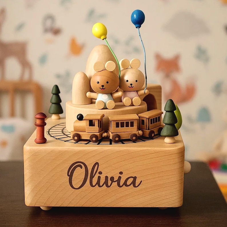 May include: Wooden music box with a train, two bear figures, and two balloons. The box is engraved with the name "Olivia". The scene includes miniature trees and a mountain-like structure, all in natural wood tones.