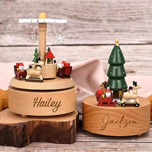 May include: Two wooden music boxes with light wood bases. The left box features a rotating carousel with a reindeer, sleighs, and snowflakes. The right box features a green Christmas tree, a reindeer, and a sleigh. The names Hailey and Jackson are engraved on the bases.