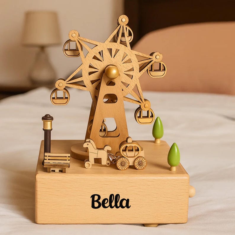 May include: A wooden music box featuring a Ferris wheel design. The Ferris wheel has small gondolas and a gold centre. The base of the box is engraved with the name "Bella". Other details include a horse-drawn carriage, trees, and a bench.