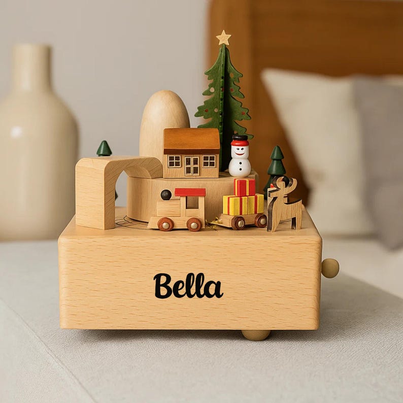 May include: A wooden music box with a Christmas theme. It features a small house, a green Christmas tree, a snowman, a train, and a reindeer. The name "Bella" is printed on the front. The box is made of light-coloured wood.