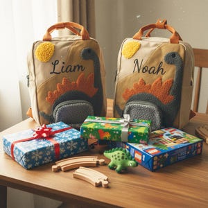 Op de afbeelding: Twee bruin en beige rugzakken met dinosaurusontwerpen en de namen "Liam" en "Noah" geborduurd. Cadeaus verpakt in blauw en groen papier, een groen dinosaurus speelgoed, houten treinrails en een Lego doos liggen op een houten oppervlak.
