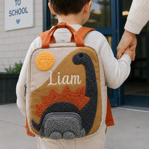 Könnte beinhalten: Ein beigefarbener Rucksack mit einer texturierten, dunkelgrauen Dinosaurier-Applikation mit rostfarbenem Stachelrücken. Der Rucksack hat eine gelbe Sonne, eine Reißverschlusstasche vorne, orangefarbene Träger und den Namen Liam in weißer Schrift.