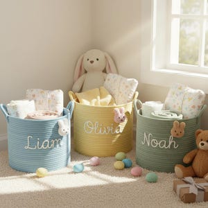 Puede incluir: Tres cestas de almacenamiento de cuerda trenzada en azul, amarillo y verde, cada una con el nombre de un niño bordado en la parte delantera. Las cestas están llenas de mantas y almohadas. Decoraciones de conejitos y huevos de Pascua están dispersos.