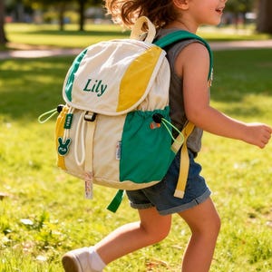 Puede incluir: Una mochila color crema con detalles verdes y amarillos. La mochila tiene el nombre "Lily" bordado en verde. La bolsa cuenta con correas ajustables, un cierre de cordón y una pequeña etiqueta con un diseño de conejo. La mochila la lleva un niño.