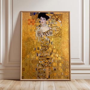 Könnte beinhalten: Gerahmter Kunstdruck eines Gemäldes von Gustav Klimt. Das Kunstwerk zeigt eine Frau in einem langen, verzierten goldenen Kleid mit geometrischen Mustern und einem schwarzen Hut. Der Hintergrund ist ein sattes Gold mit dekorativen Elementen.