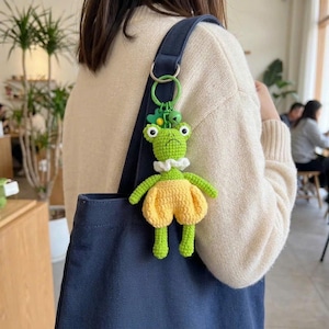 May include: A green and yellow crochet frog keychain with a white collar and yellow shorts. The frog has large, round eyes and is attached to a green metal ring. The keychain is hanging on a blue tote bag.