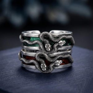 May include: Two silver rings with a snake design. Each ring has a coiled snake wrapped around a band with a green and red centre. The rings are set against a dark background.