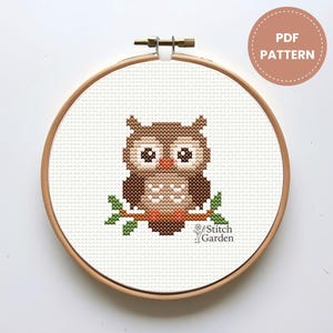 Pode incluir: Um padrão de ponto cruz de uma coruja marrom empoleirada em um galho, exibido em um bastidor de bordado de madeira. A coruja tem olhos grandes e é cercada por folhas verdes. O texto "PDF PATTERN" está em um círculo no canto superior direito.