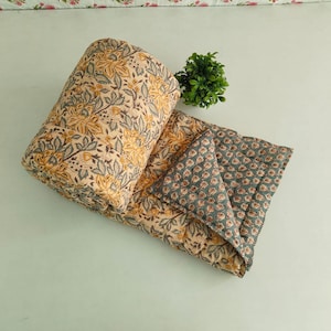 May include: A rolled-up, beige quilt with a yellow floral pattern and a gray floral patterned reverse side. The quilt is made of soft fabric and is suitable for a bed or sofa.