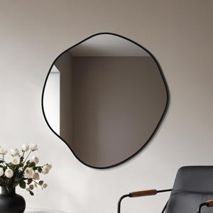 Può includere: Un interno moderno con uno specchio nero dalla forma irregolare. Sono presenti una sedia in pelle nera con braccioli in legno e un tavolino rotondo con piano in marmo e gambe nere. Un vaso nero contiene fiori bianchi.