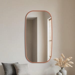 May include: A modern entryway scene featuring a large, copper-framed oval mirror. Below the mirror is a wooden bench with two gray pillows. A round, woven rug sits on the floor, and a vase with dried flowers adds a natural touch.