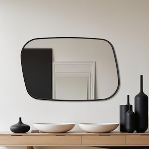 Könnte beinhalten: Ein moderner Eingangsbereich mit einem hellen Holztisch mit einem schwarzen Akzentbein. Der Tisch beherbergt schwarze Vasen, weiße Schalen und einen schwarzen, unregelmäßig geformten Spiegel. Der Raum hat ein minimalistisches Design mit einem großen Fenster.