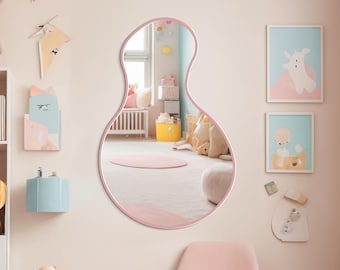 May include: A pink, organically shaped mirror hangs on a light pink wall in a child's room. The room is decorated with stuffed animals, framed artwork, and a pink chair. A white bookshelf is filled with toys and decorative items.