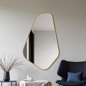 May include: An irregularly shaped mirror with a gold-colored frame hangs on a white wall. A black vase with dried plants sits on a black side table. A small wooden table with a white cloth and a blue chair are also in the room.