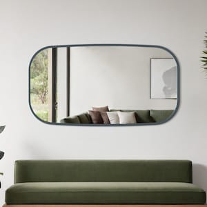 May include: A contemporary living room with a green velvet sofa, wooden frame, and a large oval mirror with a dark blue frame. Potted plants and a patterned rug complete the scene. The mirror reflects a sofa and cushions. The room has a minimalist design.
