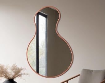 May include: An interior shot showing a copper-framed, organically shaped mirror. A black vase with dried flowers sits on a round side table. A wooden chair with a grey cushion is also present. The room has a minimalist design.