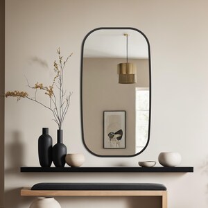 May include: A contemporary interior scene with an oval black-framed mirror. Beneath it, a black shelf displays vases and bowls. A wooden bench with a black cushion is positioned below. The color scheme is neutral, incorporating beige and black tones. The room has a minimalist design.