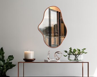 May include: An organically shaped mirror with a copper-coloured frame hangs on a light grey wall. The mirror reflects a view of a window and outdoor scenery. A modern console table with decorative items and a candle sits below.