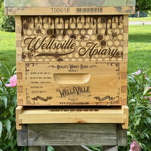 May include: A wooden beehive with the words "Wellsville Apiary" engraved on the front. The design includes bees, honeycombs, and the text "Quality Honey Bees." The hive is set on a wooden stand in a garden setting.