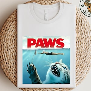 May include: White t-shirt featuring a parody of the movie "Jaws" with the word "PAWS" in red. The graphic depicts a cat attacking a swimmer in the water. A logo with a deer is in the upper right corner.