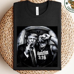 May include: Black t-shirt featuring a black and white image of two men. One man is smoking a cigar, and the other is drinking from a can. The text "Austin 3:16" is visible on the second man's tank top. The shirt has a round neck.