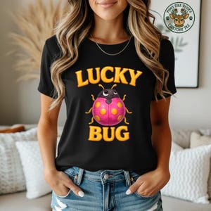 Może przedstawiać: Czarny t-shirt z żółtym napisem "LUCKY BUG" i rysunkiem biedronki z kreskówek. Biedronka ma różowy korpus z żółtymi kropkami i czarnymi czułkami. Koszulka jest noszona przez osobę.