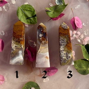 May include: Three polished stone obelisks in shades of white, brown, and grey. The stones are numbered 1, 2, and 3. The background features pink petals, green leaves, and clear crystals.