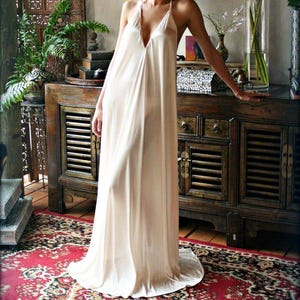 May include: A long, flowing, cream-colored halter dress with a deep V-neck. The dress is made of a soft, silky fabric and falls to the floor. The dress is worn in a room with a wooden cabinet and a patterned red rug.