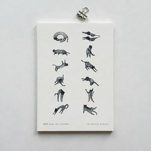 May include: A white 2027 Lazy cat calendar by Pantics Archive featuring twelve black ink illustrations of cats in various poses, secured at the top with a silver metal binder clip. The calendar displays a series of feline sketches.