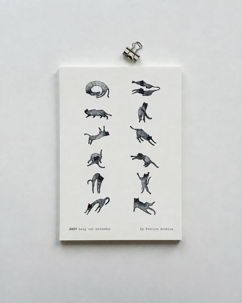 May include: A white 2027 Lazy cat calendar by Pantics Archive featuring twelve black ink illustrations of cats in various poses, secured at the top with a silver metal binder clip. The calendar displays a series of feline sketches.