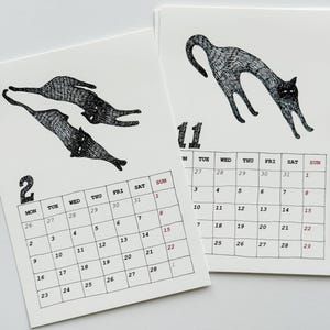 May include: Two white paper calendar pages featuring black ink illustrations of cats. The left page shows the number 2 with two cats, and the right page shows the number 11 with one arched cat. Each page includes a grid of days and dates in black ink.