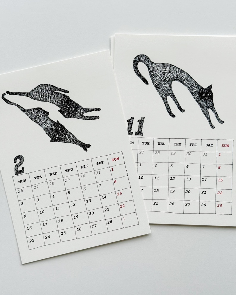 May include: Two white paper calendar pages featuring black ink illustrations of cats. The left page shows the number 2 with two cats, and the right page shows the number 11 with one arched cat. Each page includes a grid of days and dates in black ink.