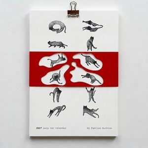 May include: A 2027 wall calendar titled 2027 Lazy cat calendar by Pantics Archive, featuring black and white illustrations of cats in various poses. A red paper band with irregular cutouts overlays the center of the white calendar page.