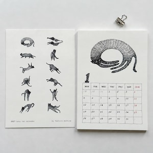 May include: A 2027 Lazy cat calendar by Pantics Archive featuring two white pages. The left page displays small black and white cat illustrations. The right page shows a large cat drawing above a monthly grid for January with the number 1.