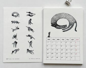 May include: A 2027 Lazy cat calendar by Pantics Archive featuring two white pages. The left page displays small black and white cat illustrations. The right page shows a large cat drawing above a monthly grid for January with the number 1.