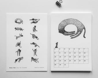 May include: A black and white calendar featuring whimsical illustrations of cats in various poses. The calendar includes a monthly layout with dates and the text "Every Year Lazy Cat Calendar" and "Pantics Archive". A binder clip is at the top.