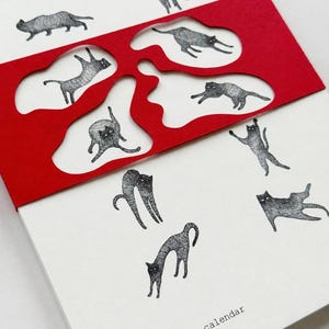 May include: A 2027 wall calendar featuring black and white ink illustrations of cats in various poses. A bright red paper overlay with organic, irregular cutouts partially covers the white pages, revealing some of the cat drawings underneath.