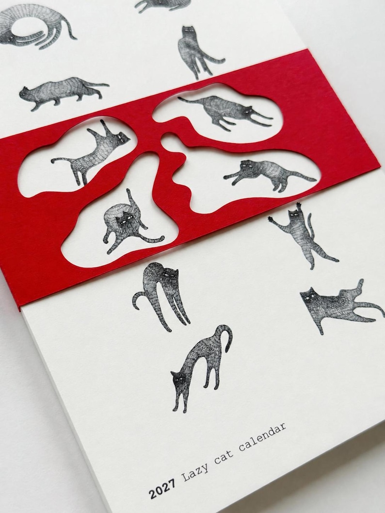 May include: A 2027 wall calendar featuring black and white ink illustrations of cats in various poses. A bright red paper overlay with organic, irregular cutouts partially covers the white pages, revealing some of the cat drawings underneath.