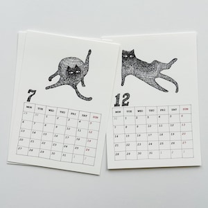 May include: Two white calendar pages featuring black ink illustrations of cats. The left page shows the number 7 with a cat grooming itself, while the right page shows the number 12 with a cat stretching. Each page includes a grid of days.
