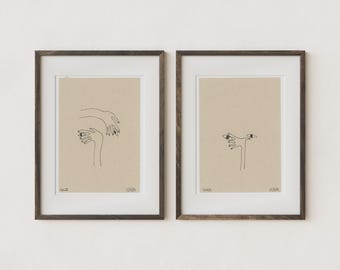 May include: Two framed art prints featuring minimalist line drawings of hands and eyes. The artwork is in black ink on a tan background, set within a white mat and dark brown wooden frames. The prints are displayed on a white wall.