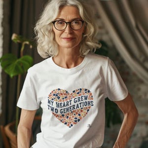 May include: White t-shirt featuring a heart design composed of paw prints and leaves in blue, orange, and brown. The text "MY HEART GREW TWO GENERATIONS" is within the heart. The person is wearing glasses.