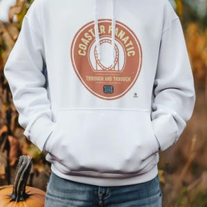 May include: A white hooded sweatshirt featuring a circular graphic on the chest. The graphic contains the text COASTER FANATIC, an illustration of a roller coaster loop, the text THROUGH AND THROUGH, and a small ticket icon at the bottom.