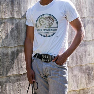 May include: White t-shirt with a circular graphic that reads "River Bass Hunter at Heart" and a bass fish illustration. The person is wearing a black belt with a "Beatles" buckle and gray jeans. They are holding binoculars.