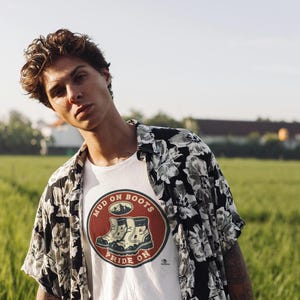 Op de afbeelding: Een persoon draagt een wit t-shirt met een cirkelvormige afbeelding met de tekst "MUD ON BOOTS PRIDE ON" en een zwart-wit overhemd met bloemenprint. De achtergrond is een groen veld en een blauwe lucht.