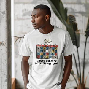 以下が含まれることがあります： カラフルなカレンダーのグラフィックと「I MISS SILENCE BETWEEN MEETINGS」のテキストが入った白いTシャツ。カレンダーには様々な模様の正方形があり、その上に雲があります。Tシャツは半袖です。