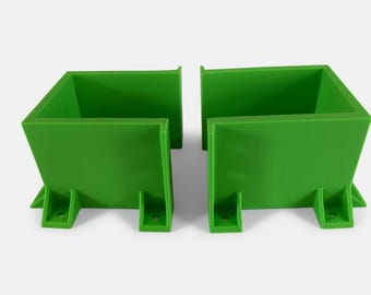 Puede incluir: Dos contenedores verdes brillantes, de forma cuadrada, con una base trapezoidal y una abertura rectangular. Cada contenedor tiene dos pequeños soportes rectangulares con agujeros en la parte inferior. Los contenedores están colocados uno al lado del otro.